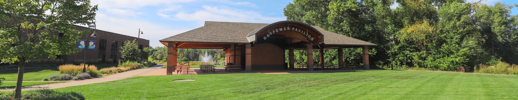 Wide shot of a large pavilion. Freshly mowed grass is in the foreground. Gardens and a water fountain can be seen in the background.